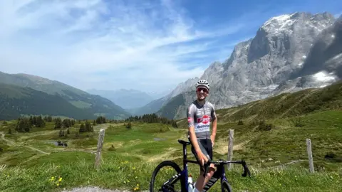Supplied Jente Michels is smiling at the camera in a helmet and cycling gear. He is posing on his bike with mountain slopes towering behind him.