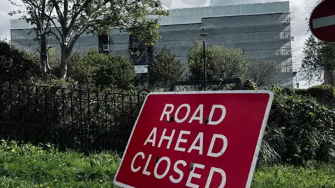 This image is taken from lower down the road and puts a large red warning sign in the foreground. The sign reads “Road ahead closed” in bold white capital letters and is propped up on a black metal stand on a patch of grass at the roadside. Behind it, there is a low black iron fence and dense greenery, including bushes and small trees, which partly obscure the view beyond.