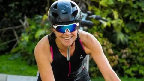 Rebecca Comins cycling and grinning with a bike helmet and sunglasses on 