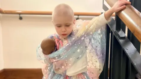 HANDOUT A little girl with very short hair walking down some stairs wearing a sparkly dress and holding a doll.