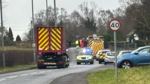A road with a lorry, a police car with lights flashing, a fire ambulance and several other cars on it. People in high viz clothing are in the distance. On either side of the road there are circular 40 miles per hour signs.