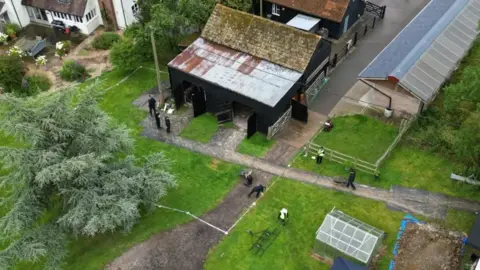 Steve Hubbard/BBC Aerial view of Stocking Farm as police search for body of Muriel McKay in July 2024. Foreground shows a tall fir tree surrounded by grass. Behind it a barn building with black walls and a metal roof, and a simlar building behind that. Right foreground there is a greenhouse and next to that a dug up patch of soil. Police officers are seen from above wearing black clothing