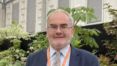 A man in a shirt, blaze and tie standing outdoors in front of plants. He has an orange lanyard around his neck and is smiling at the camera. 