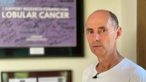 Peter Whittlesea / BBC A man stood in front of a framed purple sign reading "lobular cancer".