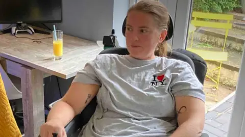 Stacey is sat in an adapted chair in a grey t'shirt and her brown hair tied back. She looks to be in the kitchen of her home next to a table and chair with glass doors to her garden behind her.