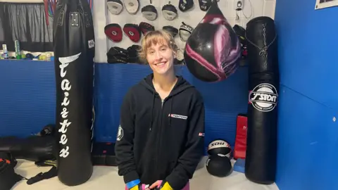 Miriam Grubb stood between two black punch bags which has writing on it. She is wearing a black jumper with her hands in front of her. On the walls there is blue padding and gloves hung up above them. 