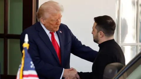 Trump and Zelensky shake hands at the White House after the Ukrainian president gets out of his car on arrival. Trump is wearing a dark blue suit and red tie, Zelensky in a black military-style outfit.