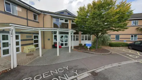 Google Maps The front of a two-storey hospital building with light orange bricks. It has white pillars holding up a glass roof above its pavement. Emergency and disabled parking bays can be seen in the foreground.