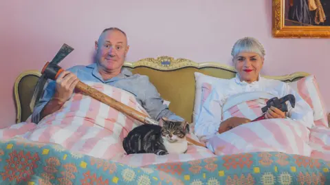 Keith and Marj are sat up in a bed with pink and white striped bedding. There is a cat sat between them. Both of them are holding building tools. The wall behind them is pink and has a hole in it exposing wooden panels. On the same wall is a gold mirror.