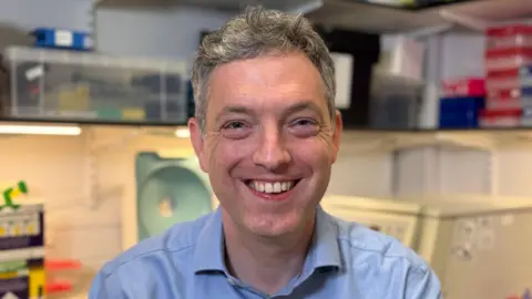 Consultant haematologist Dr Ben Uttenthal in a room at Addenbrooke's Hospital. He has short grey hair, a light blue shirt and is smiling into the camera. Blurred out behind him are clear plastic boxes containing equipment.