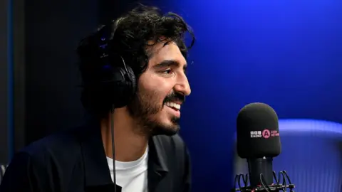 BBC Asian Network Dev Patel in the BBC Asian Network studio. Dev is a 33-year-old British Asian man with dark curly hair, a moustache and short beard and brown eyes. He’s photographed from the side, smiling. He is pictured inside the studio, which has blue walls, and wears headphones as he sits in front of a microphone. He wears an unbuttoned black shirt over a white T-shirt