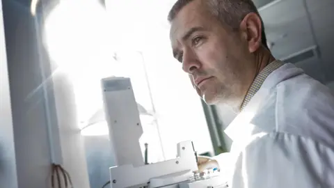 Dr Stephen Grimes, Associate Professor in Stable Isotope Geochemistry and Climate Change at the University of Plymouth. He is stood up and looking to the left. He is wearing a white lab coat. In front of him is a microscope and bright light. 