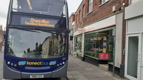 Go East Anglia A blue and grey Konectbus. It is parked outside a shop and on the front of the bus it says "Chatty Bus tackling loneliness".