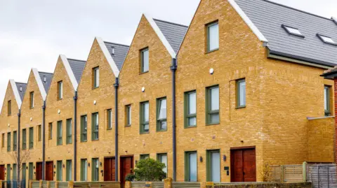 A row of six red brick houses with red wooden doors. Each house is identical with one window on the 2nd floor, three on the first floor and two to the left of the door on the ground floor. 
