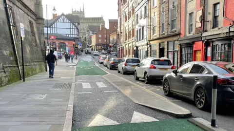 A green cycle lane with a pedestrian crossing in the middle leading to a narrow bus stop. Cars stuck in traffic can be seen next to the bus stop. Pedestrians are walking on the pathway to the left of the image.