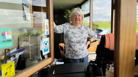 Woman in a blue stripy top stands next to an open hatch/window. A set of scales is on one side and the green countryside can be seen through a window behind her. 