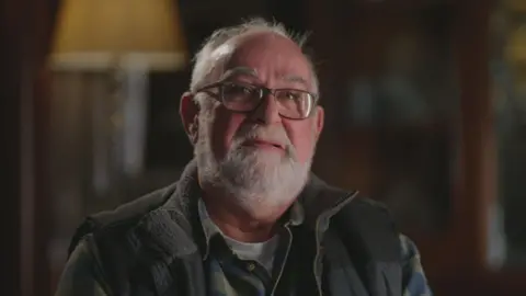 Journalist Andy Spearman looks into the camera done  his glasses successful  a acheronian  lit room, with the inheritance  retired  of focus. He has grey thinning hairsbreadth  and a abbreviated  grey beard. Andy is wearing a achromatic  t-shirt, greenish  and navy checked garment  and a navy assemblage  warmer. 