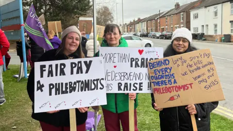 Three phlebotomists - all women dressed for cold weather - stand on a grass verge at the side of a road as part of a picket line. They are all holding signs on sticks which read slogans such as "Fair pay for phlebotomists" and "behind every diagnosis, there is a phlebotomist". Other people taking part in the strike can be seen behind them.