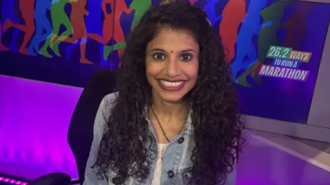 BBC Madhusmita Jena, who is smiling at the camera, is sitting in the BBC Radio Manchester studio with a sign behind her which reads: 26.2 Ways To Run A Marathon.