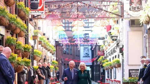 PA Media King Charles and Queen Camilla on a cobbled street in Belfast. Above them are different coloured umbrellas hanging on lines. The King is wearing a blue suit with a light shirt and blue polka dot tie. The Queen is wearing a green coat and dress with boots. 