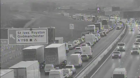 National Highways A black and white CCTV image showing the A14. The left lane is blocked by traffic. There is a large sign to the left of the road pointing left for St Ives, Royston and Godmanchester. 