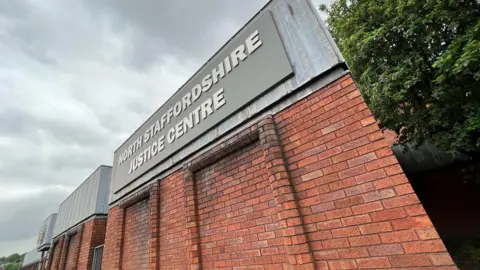 A brick building with a metal band around the top and a grey sign which reads North Staffordshire Justice Centre in white writing.