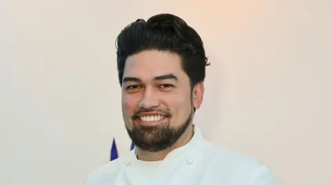 Luke Selby, wearing a chef's white outfit, smiles at the camera. He has dark hair and a short dark beard