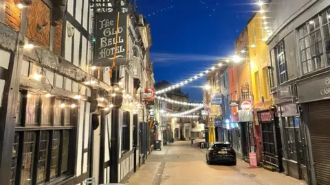 A winding street with shops and pubs either side, with a zig-zag of lighting strung across the street about two-storeys high.