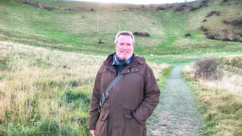 James Neale at the Long Man of Wilmington. James is wearing a brown coat and is carrying a bag. He has graying hair and is smiling to camera.