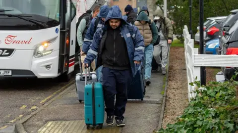 A group of men, wearing hooded jackets, walk along a street dragging suitcases after disembarking from a white coach.