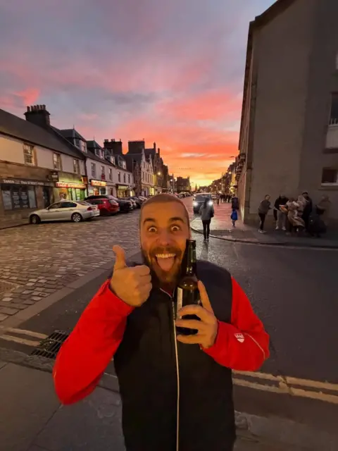 Justin Bieber Justin Bieber gives a thumbs up sign while holding a beer outside. A cobbled street and shops are seen in the background