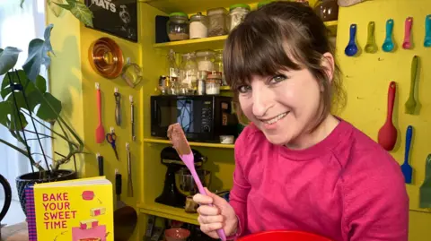 Tat Effby A woman with short brown hair, a pink top and a pink spatula in a room with yellow walls, beside a yellow book