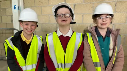 Three children smile at the camera - they are nine and ten years old. Two boys on girl. They are all wearing yellow high-vis vests and white hard hats. They stand in front of a wall.