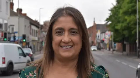 Head and shoulders of Nisha Popat smiling wearing a green flowery top. There is a road with cars in the background.