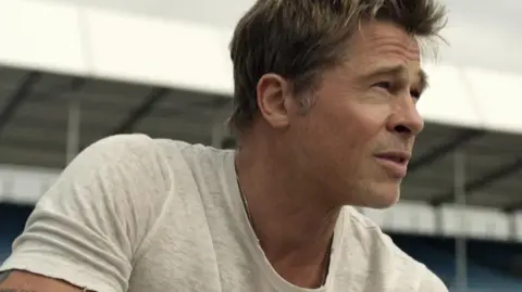 A still of the F1 trailer with Brad Pitt in a beige T-shirt looking into the distance