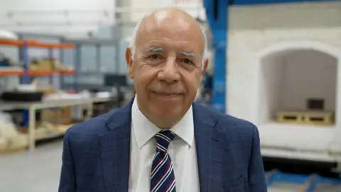 A bald man wearing a white shirt and navy blue suit jacket with a blue, white and red striped tie is standing in what appears to be a factory. The room is brightly lit, and there are work benches and racking behind the man.