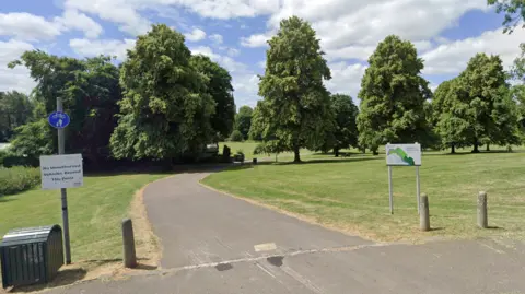The entrance to Monkton Park as seen from an adjacent road. There is a winding tarmac path going into the park, which is broad and grassy, with neatly-space tall leafy trees in the background.
