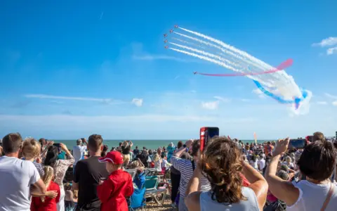 Getty Images Crowds watching the RAF Red Arrows aerobatic display team from the beach at previous airshow in Eastbourne. There are six aircraft in view, with red, white and blue vapour trails visible in the sky.
