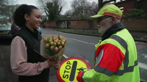 A student in a gilet and pink sweatshirt hands a bunch of flowers to John at the side of a road outside Sale Grammar School. 