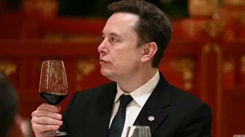 Musk, wearing a black suit, white shirt and black and white dotted tie, holds a glass of red wine while at an event in Beijing, during a recent US trade visit.