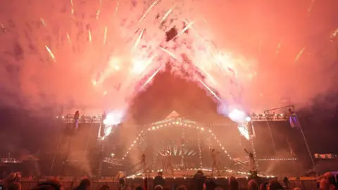 Acrobats and circus performers take part in the opening ceremony in the Pyramid Arena at Glastonbury Festival. There are red fireworks going off around the stage.