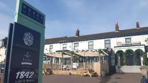 The Roker Hotel is a two-storey white building with an arched entrance and a seating area outside. A tall blue sign outside reads Roker Hotel.