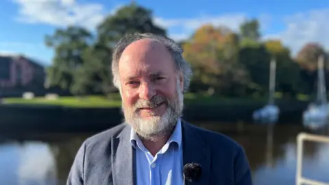 Julian Brazil pictured by a body of water wearing a navy blue jacket and light blue shirt. He has a grey and white beard with short receding hair. 