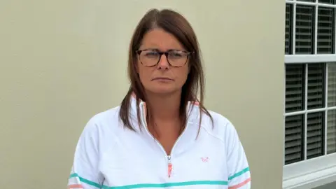 Melissa has dark shoulder length hair, and glasses, and wears a white quarter zip top. She stands in front of a beige coloured outside wall of a house with a window to her right. 
