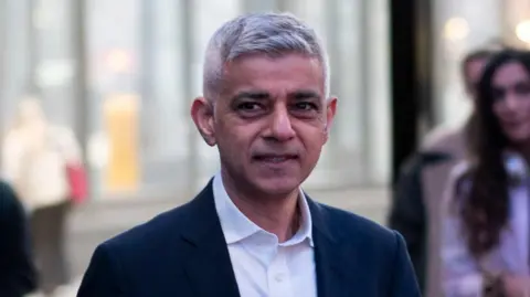 PA Media London mayor Sadiq Khan is wearing a dark suit jacket and a white shirt. There are people standing to the side taking photos of him.