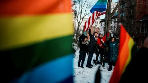 An out of focus rainbow Pride flag flies in the foreground as a small group of people raise their arms in celebration at the Stonewall Monument site. 