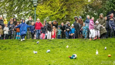 Micheal Porter Photography A group of people wearing coats and winter clothing stand at the top of a grassy hill, with large, designed eggs rolling down it. Some of the eggs are in plastic bags. There are leafy trees and a lamppost in the background.