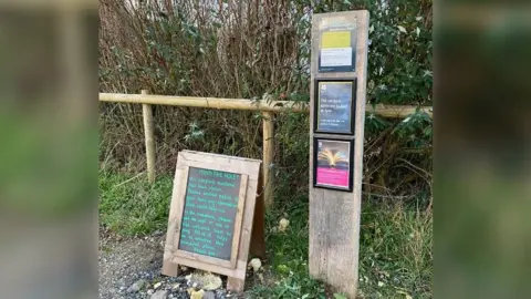 A sandwich board sign stands next to a tall, skinny wooden plaque, with framed posters on it, at the edge of a car park in the countryside. The sandwich board sign reads, in green chalk, "Mind the hole! Our carpark machine has been stolen. Please contact police if you have any information that could help us. In the meantime, please use the cafe or one of the welcome team to pay. All of it helps us to conserve this beautiful place. Thank you!".