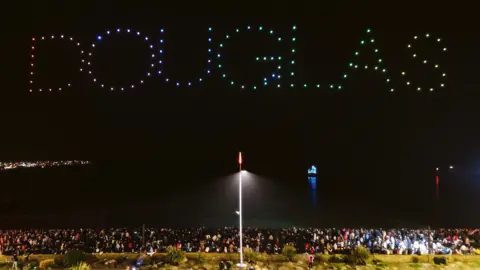 Douglas Council/Matt Mosur Drone lights spelling Douglas over crowds watching from the promenade. There is one street light on in the centre of the frame.