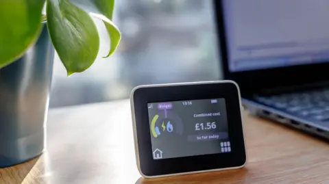 Getty Images a domestic energy smart meter showing a daily cost of £1.56 is sitting on a table with a laptop computer and a potted plant beside it